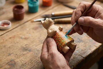 A craftsman's hands carefully painting a colorful pattern on the skirt of a carved wooden cat figurine.