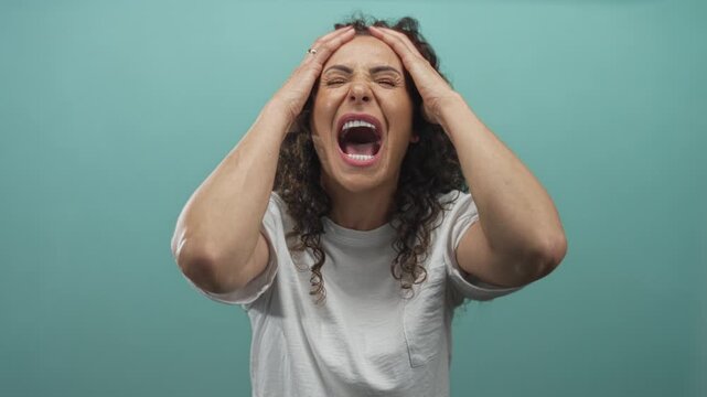Young hispanic woman screaming with mouth wide open in a mint green studio setting; frustration.