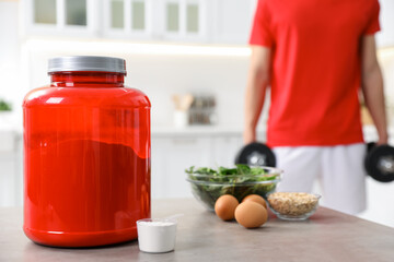 Man with dumbbells in kitchen, focus on protein powder in measuring scoop, jar and ingredients