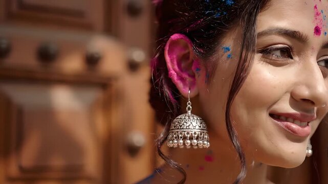 Smiling young Indian woman, face and ear adorned with vibrant pink and blue Holi colors, wearing traditional jhumka earrings. Perfect for celebrating Indian festivals, cultural traditions, joy,
