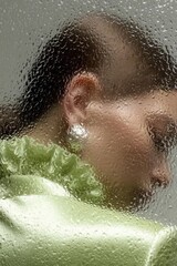 Artistic side portrait of a woman in a green outfit seen through textured glass, creating a soft, dreamy effect that expresses elegance and introspection.