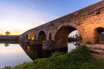 Fototapeta premium Romantic twilight photograph of the ancient Puente Romano arched roman bridge over the Guadiana River in Merida Spain (ESP) with vibrant sunset reflections