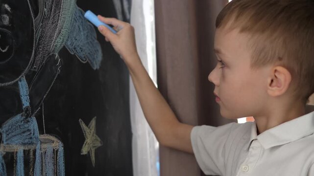 Focused little boy drawing with blue chalk on a blackboard at home or in classroom. Early education, creativity and childhood development concept. Side view portrait with copy space