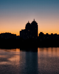 Fototapeta premium Urban towers silhouette against sunset sky. Modern city buildings with spires against a glowing orange and blue sunset, soft reflections on the water in the foreground during twilight.