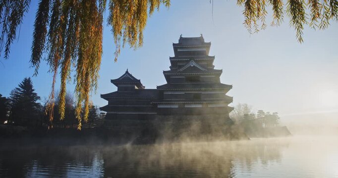 Cinematic silhouette of 16th century Matsumoto castle with misty pond and autumn weeping tree