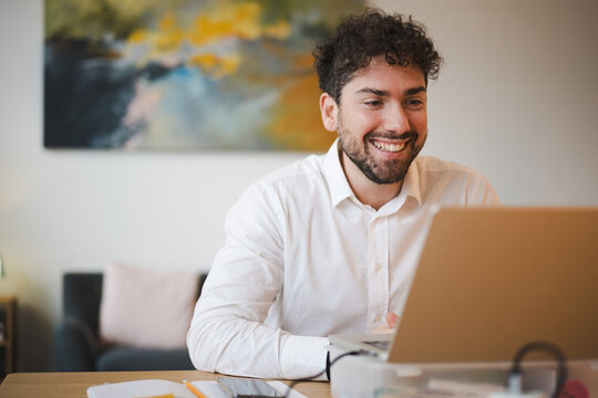 A smiling man in a white shirt works on his laptop at a desk, with a phone and notebook nearby.