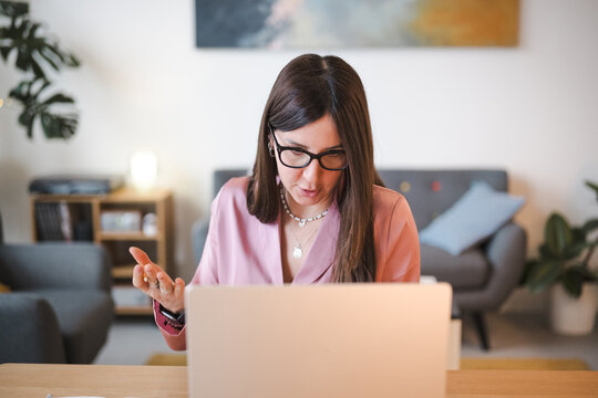 A woman wearing glasses and a pink blazer is actively engaged in a video call on her laptop, gesturing with her hands as she communicates.