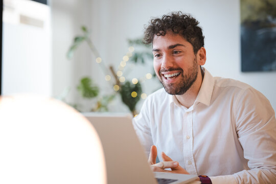 A man with curly hair smiles while looking at his laptop. He is wearing a white collared shirt and has his hands clasped.