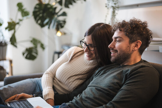 A young couple smiles while looking at a laptop together on a couch, enjoying a shared moment of connection and entertainment in a cozy home environment.