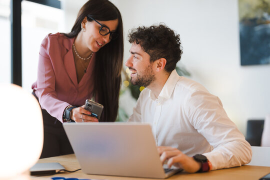 A woman shows a man something on her phone while he works on a laptop, collaborating in a modern office setting.