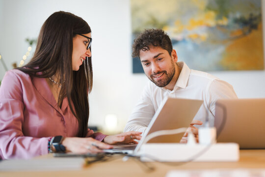 A man and a woman collaborate on a project, looking at a laptop screen together. They are seated at a table in a modern office setting.