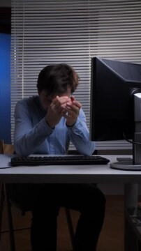 Vertical video overworked man at a computer desk feeling overwhelmed and frustrated, holding his head in his hands, reflecting the stress of a demanding job and late night work environment