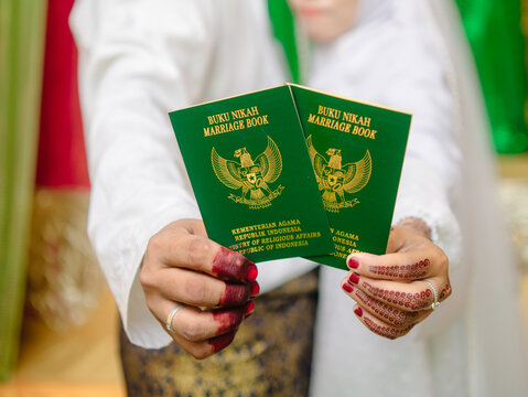 Newlywed couple hands holding Indonesian green marriage books. Close up of bride with henna pattern and groom in white shirt. Perfect for wedding concept shots.
