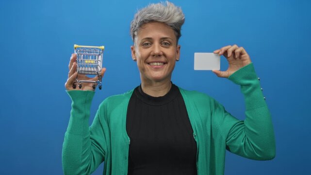 Woman wearing green cardigan holds miniature shopping cart and credit card in studio; financial literacy confidence.
