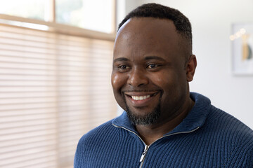 African American man smiling in soft light near blinds, framed artwork, lights, in blue zip sweater © wavebreak3