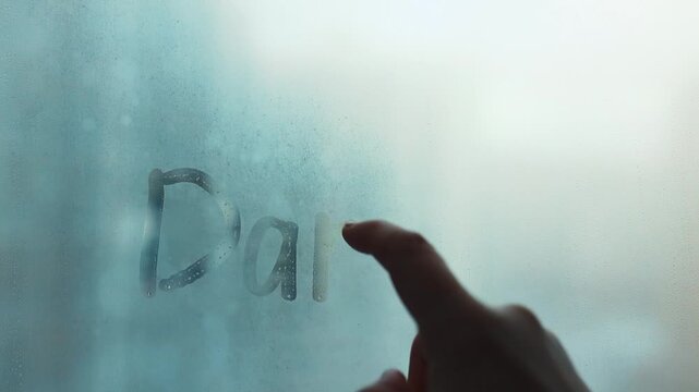 Female hand is writing lettering German word Danke is Thanks in English on misted rainy glass on blue window with raindrops