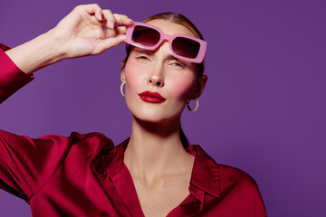 Stylish woman in red satin blouse adjusts pink sunglasses on purple background. Fashion portrait with glossy red lips and hoop earrings. Concept of glamour, confidence, and modern style. © SHOTPRIME STUDIO