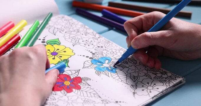 Woman coloring antistress picture with felt tip pen at light blue wooden table, closeup