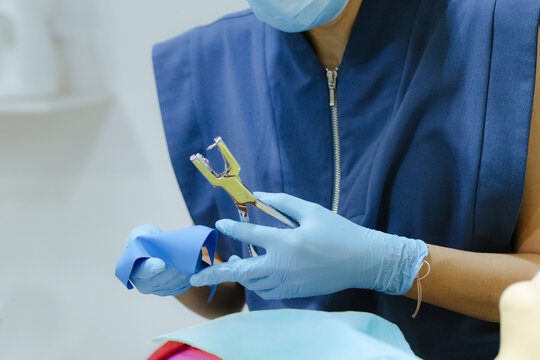 Dental professional in blue scrubs and gloves preparing a rubber dam with a puncher for an upcoming dental procedure