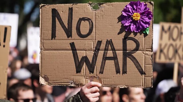 A hand holds up a cardboard protest sign that reads "No WAR" in bold, black letters.
