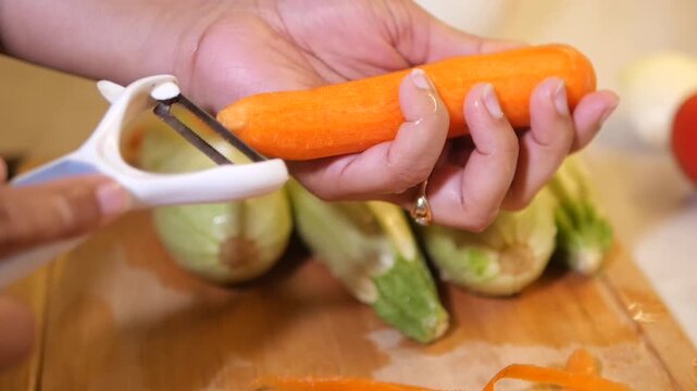 Woman peeling fresh orange carrot with white vegetable peeler over wooden cutting board