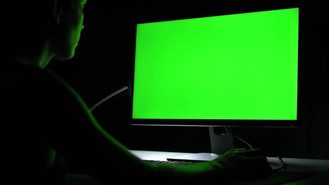 A person working on a computer with a green screen monitor in a dark room, viewed from the side, displaying a blank green screen.
