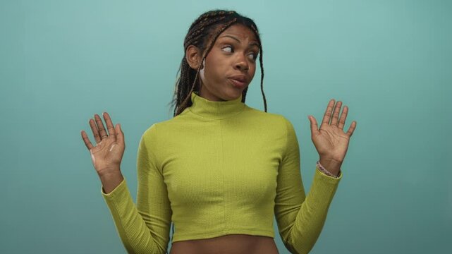 Woman raising hands with visible palms and nose piercing in teal studio wearing lime turtleneck and braids; dismissal shrug.
