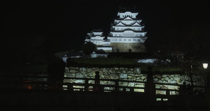 Silhouette of bridge near glowing stone wall and bright 16th century Matsumoto castle - wide shot at night