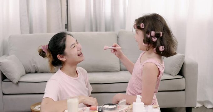 Multiracial girl with hair rollers applying makeup to Asian woman sitting on floor together smiling in light living room surrounded by cosmetic bottles and glass of milk