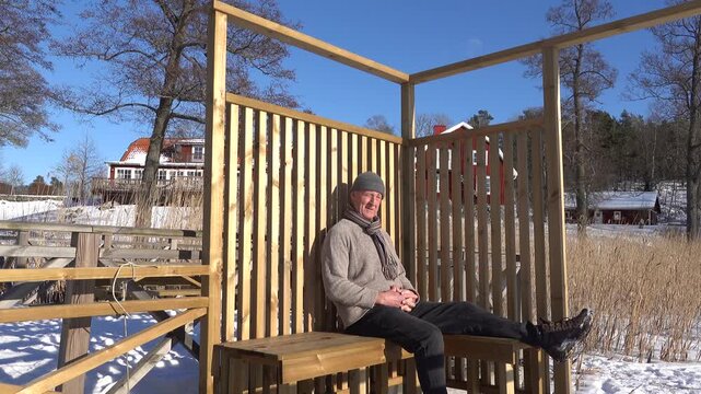 Trosa, Sweden A man sits in the winter sun on a wooden sundeck by a frozen lake. 