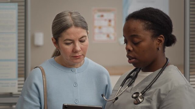 Female doctor and patient of different ethnicities analyzing digital medical test results together on tablet during in depth consultation in modern clinic environment