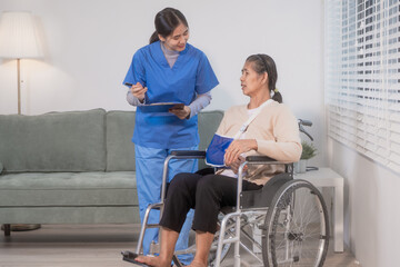 Fototapeta premium A young Asian nurse discusses medical information with an elderly female patient seated in a wheelchair with an arm sling at home, highlighting professional healthcare support, rehabilitation