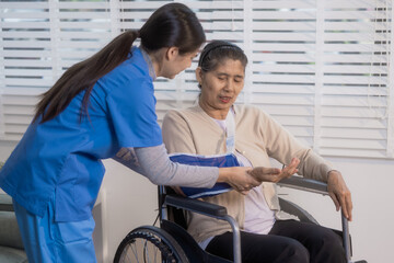 Obraz premium A young Asian nurse discusses medical information with an elderly female patient seated in a wheelchair with an arm sling at home, highlighting professional healthcare support, rehabilitation