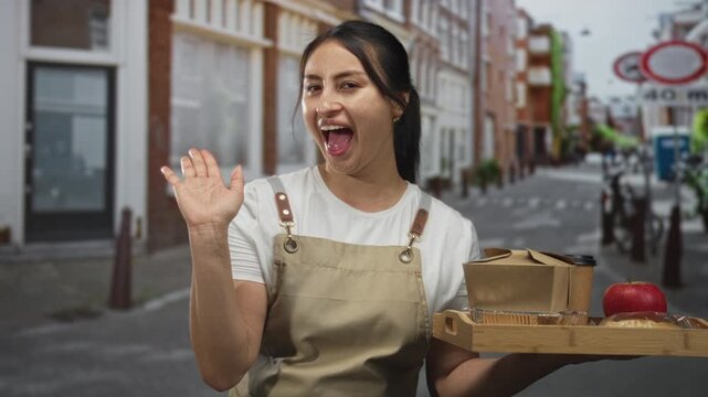 Young hispanic woman in apron holds wooden tray with takeaway box apple and blows kiss while waving on street; friendly service.