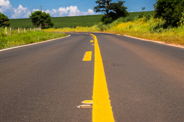 Asphalt road with yellow lines curves through the vibrant green landscape of rural Brazil under a blue sky. A concept of journey, travel, and perspective in South America's countryside.