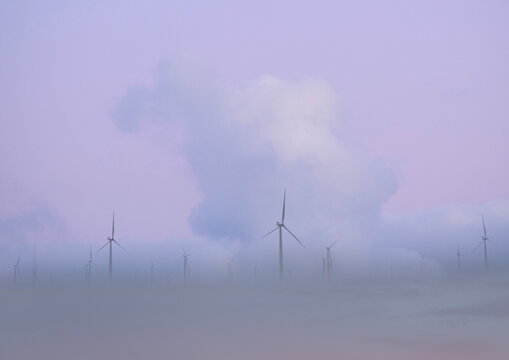Pink and purple sky, cloudy and misty with wind turbines towering above the clouds.
