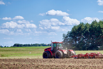 Fototapeta premium Red tractor cultivating agricultural field, preparing soil for sowing crops, farming industry working in rural landscape