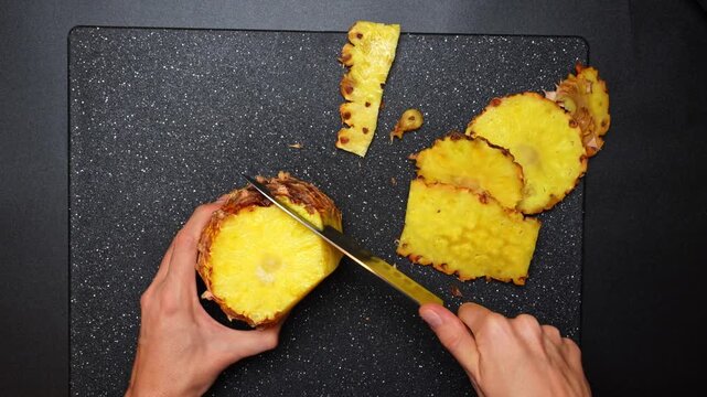 Person slicing fresh pineapple on a dark board. Top view of hands using a knife to cut the skin off a fresh yellow pineapple on a black surface.