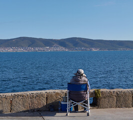 Fototapeta premium Individual seated on a folding chair by the waterfront, overlooking calm blue waters and distant hills under a clear sky