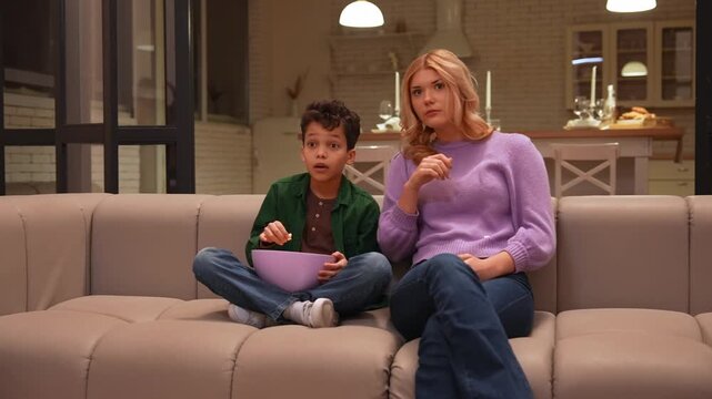 Responsible pretty teen nanny protecting adorable African American school age boy with popcorn from adult content on tv, covering child eyes with hand while watching movie in evening in domestic room.