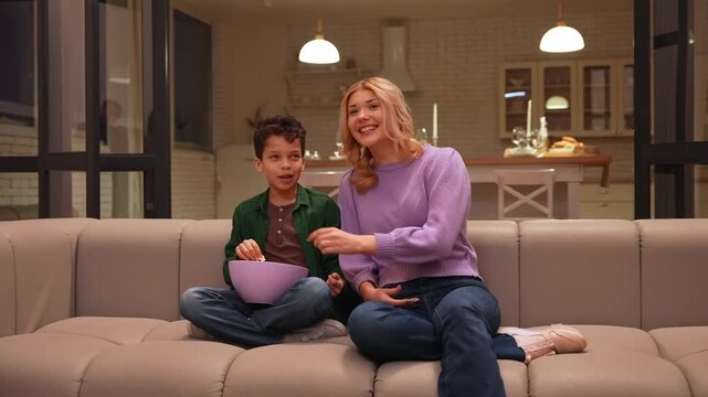 Laughing attractive teenage babysitter and joyful school age black kid with popcorn enjoying leisure , watching comedy movie on subscription based streaming service in evening in domestic room.