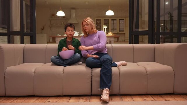 Portrait of pretty teen babysitter and cute preadolescent black boy with popcorn sitting on sofa, watching horror movie on tv, expressing fear and shock in evening in domestic room