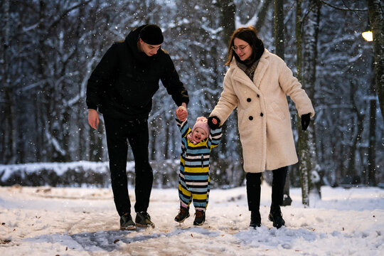 Parents walk their toddler along snowy winter park path in evening.