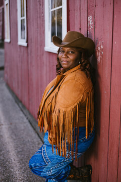Adult black woman leaning on red wooden barn in cowgirl clothes