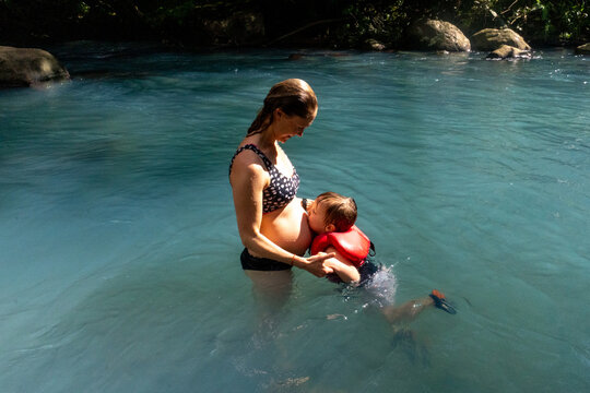 A toddler kisses his mother's pregnant belly while swimming.