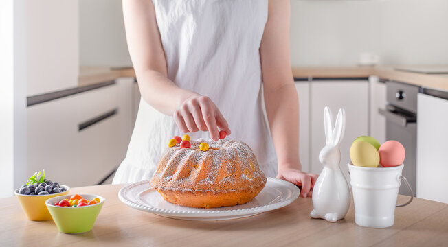 Easter cake being decorated in the kitchen. A person decorating a traditional Easter bundt cake in a bright kitchen setting. The scene includes pastel‑colored eggs, a ceramic bunny, and fresh fruit, c