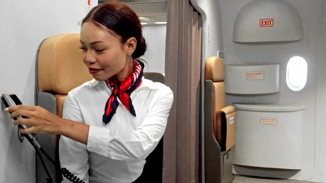 Flight attendant on a jumpseat uses the intercom for a safety announcement near an aircraft exit door, smiling in uniform during airline operations.
