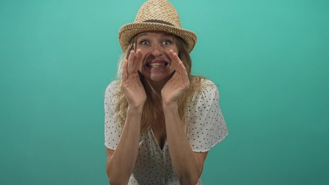 Woman cupping hands around mouth and calling out in studio with teal backdrop, wearing straw hat and polka dot dress; playful secret.