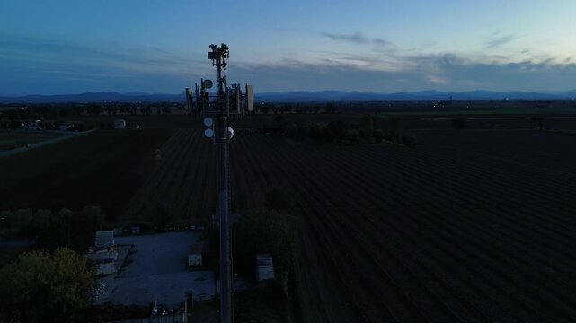 Aerial footage of a tall telecommunication tower equipped with antennas and satellite dishes, transmitting signals against a clear dark blue sky at dusk in a rural landscape
