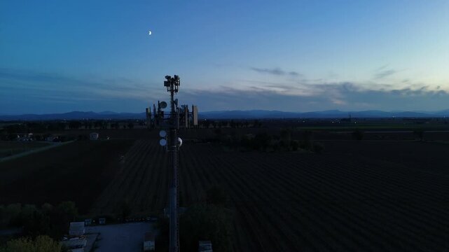 Aerial footage of a tall telecommunication tower equipped with antennas and satellite dishes, transmitting signals against a clear dark blue sky at dusk in a rural landscape